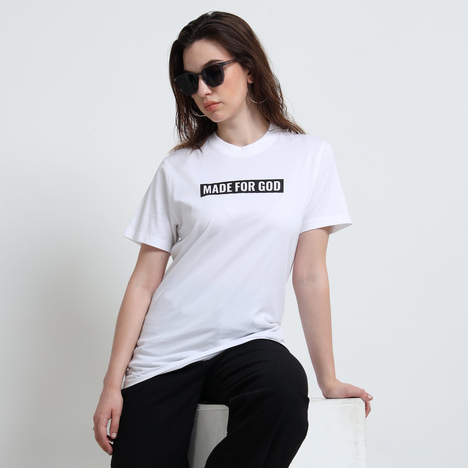 Person wearing a white t-shirt with 'MADE FOR GOD' text, sitting on a white block against a white background.